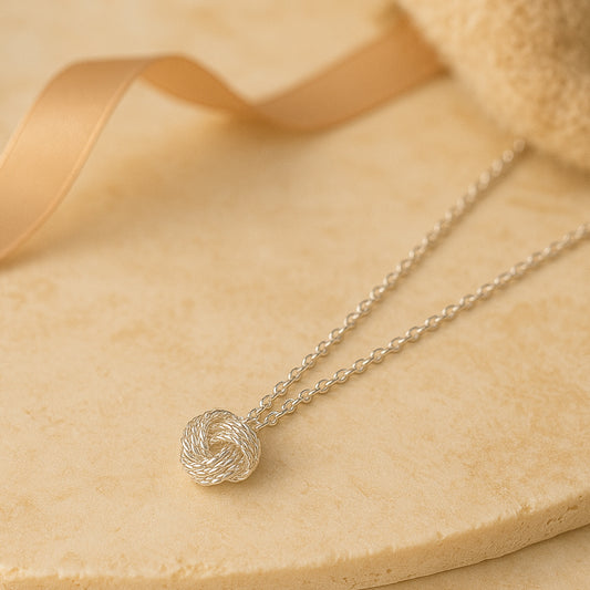 Feine silberne Halskette mit elegantem Kordelknoten-Anhänger auf beigem Untergrund - stilvoller und zeitloser Damenschmuck.